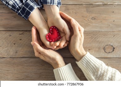 Red heart in child and female hands over wooden background, copy space. Kindness, family, love and charity concept, hand made valentine or mother's day gift.