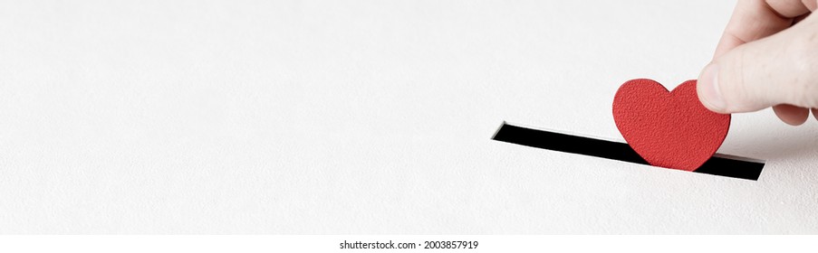 Red heart symbol is put by person's hand into slot of white donation box. Concept of donorship, life saving or charity, sincere devotion to faith. Close-up shot, horizontal banner with copy space - Shutterstock ID 2003857919