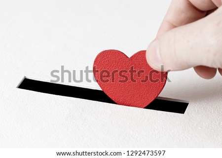 Red heart symbol is put by person's hand into slot of white donation box. Concept of sincere devotion to faith. Concept of donorship, life saving or charity. Close-up shot
