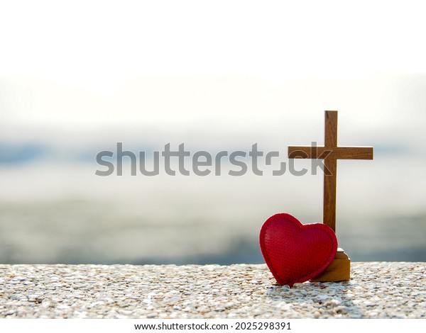 Red heart with wooden Christian cross on gravel\
floor in morning light, beach sea as background. Jesus love you.\
Faith hope believe in God. Believe in salvation. Christianity\
background concept.