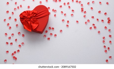 Red heart-shaped gift box with a red ribbon on the left side on a white background with little red crystals spilled around. Happy Valentine's Day. Top view flat lay copy space.