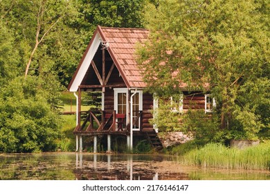Red holiday cabin with reflection in the pond. Wooden cottage, sauna on shore. Tiny house near the water. Building surrounded by green trees.