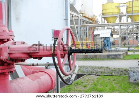 Red hydrant fire fighting valve dominates foreground with industrial pipes. Background shows factory structures and yellow silos. Green grass and concrete frame the scene. Industrial safety equipment is clearly visible
