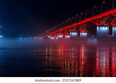 A red illuminated bridge stretches over a calm river at night, with mist floating above the water and colorful reflections. The serene urban scene captures the structure and lighting - Powered by Shutterstock