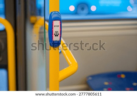 Red illuminated stop button mounted on a yellow pole inside a public bus with blurred seats and windows in the background for passenger use.