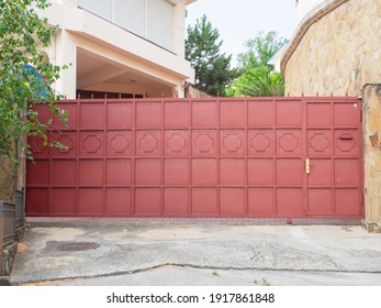 A red iron fence with a wicket gate, geometric pattern and sharp spiers encloses a modern residential building and a courtyard with various green trees