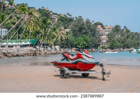 Red jet ski on trailer stands on sand beach, seashore and palm trees, morning time, tropical resort, Thailand. Front side view