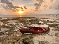 A Red Kayak Rests On Ocean Washed Rocks As The Tide Slips Away During A Warm Sunset With A Long Bridge In The Distance At Bahia Honda Key, Florida.