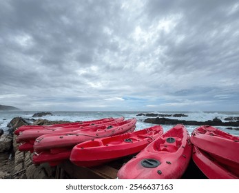 Red kayaks stacked at the coast. Ocean waves crashing against the rocks, overcast sky, cloudy day by the sea - Powered by Shutterstock