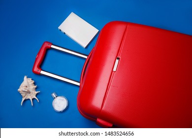 A red large travel suitcase on wheels with a retractable handle, surrounded by a perfume bottle, seashell and a white blank book. Blue background. Travel concept.
