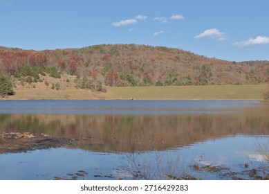 red leafed trees and bare fall tree covered hills reflecting on a lake. the peaceful reflection of the hills on a manmade lake