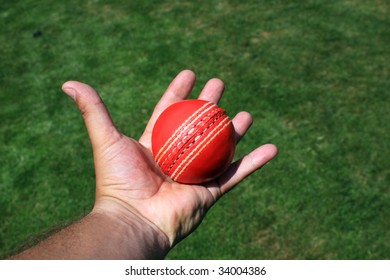 A Red Leather Cricket Ball Caught In An Open Hand. Set Over A Green Grass Area.