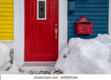 A Red Letterbox Or Mailbox On A Vintage Blue Wood House With A Red Metal House Door. The Adjoining Exterior Wall Of The Red Door Is Bright Yellow. There's White Snow Piled In Front Of The Blue Wall.