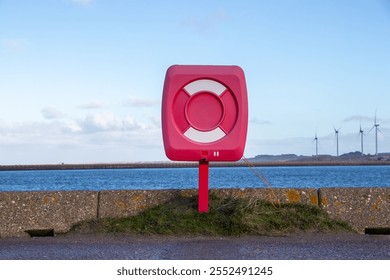 A red lifebuoy station mounted on a post near the water, with wind turbines visible in the background, set against a blue sky and coastal scenery. - Powered by Shutterstock