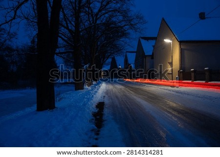 Red Light Trails Cut Through a Snowy Village Street as Winter Evening Settles Over the Countryside - A quiet village street covered with snow is illuminated by warm streetlights during a cold winter evening. Long-exposure photography transforms the passing vehicle into vivid red light trails that streak through the scene, creating a striking contrast against the blue tones of dusk. Snow-covered rooftops, bare roadside trees, and reflective pavement combine to create a visually rich composition that balances motion and stillness. The image captures the atmosphere of rural life after sunset, when artificial lighting begins to dominate the landscape and occasional traffic leaves temporary traces in the darkness. The photograph conveys themes of winter travel, transportation, village life, seasonal weather, and the contrast between human activity and the calm of a snowy night.