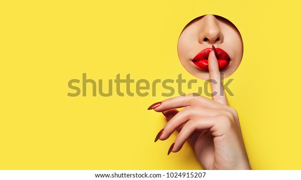 Red lips look through the hole in the yellow\
paper. A finger with a red varnish at the mouth.Fashion, beauty,\
make-up, cosmetics, beauty salon, style, personal care, geometry,\
texture, bright.