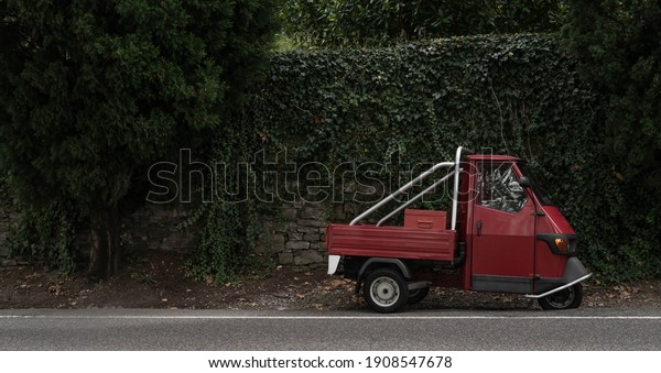 Red, little retro car or delivery truck on the\
road among green wall of plants. Typical red and green italian\
picture. in dark, dull\
colors.