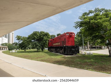 Una locomotora roja exhibida al aire libre en un área cubierta de hierba rodeada de árboles en el parque La Plancha, Mérida, México, con una pasarela de hormigón en primer plano y un cielo azul claro por encima.
