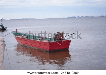 A red long-tail boat with an old engine and ropes rests calmly on the still water, reflecting a simple yet practical fishing vessel.