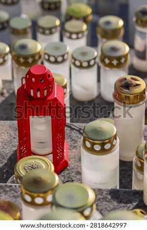 A red memorial lantern and many various plastic and glass grave candles have been placed on the stone steps of the cemetery’s memorial site. Small focus area.