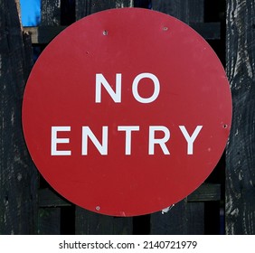 A red metal circular sign on a black painted wooden plank fence says ‘no entry’. A square of blue sky can be seen through a gap between the planks. - Shutterstock ID 2140721979