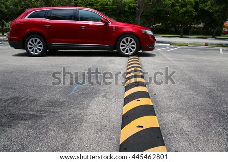 Red Minivan Driving Up to and Just Connecting with Yellow and Black Striped Speed Bump in Parking Lot with Diagonal Striped Spaces and Trees in the Background, Mid-Day