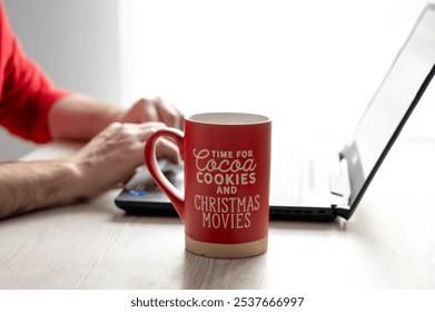 Red mug with festive text next to a person working on a laptop, capturing a cozy, holiday-themed workspace atmosphere - Powered by Shutterstock