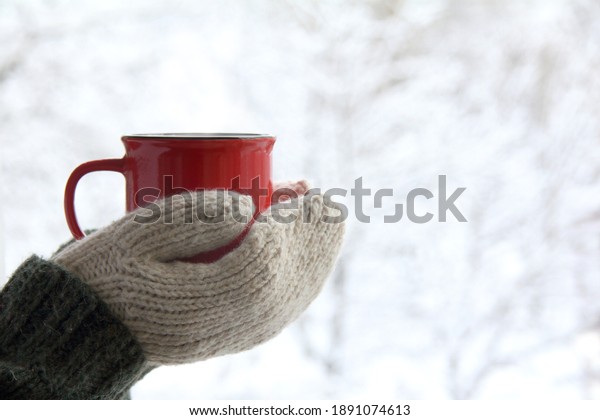 a red mug in the hands of those dressed in\
knitted mittens against the backdrop of a blurred snow landscape. a\
warming drink for a winter\
morning