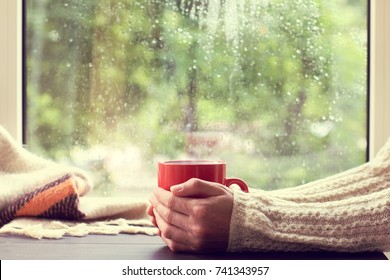 Red Mug Of Hot Drink In Hand, When Behind Is Window Is Rain / Cozy Home Atmosphere In Autumn