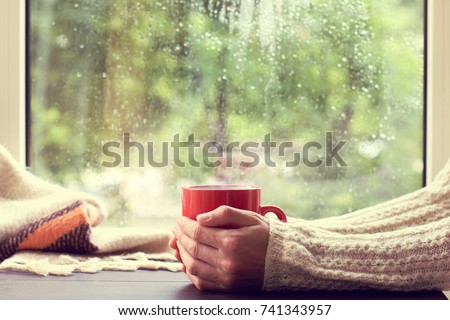 red mug of hot drink in hand, when behind is window is rain / cozy home atmosphere in autumn