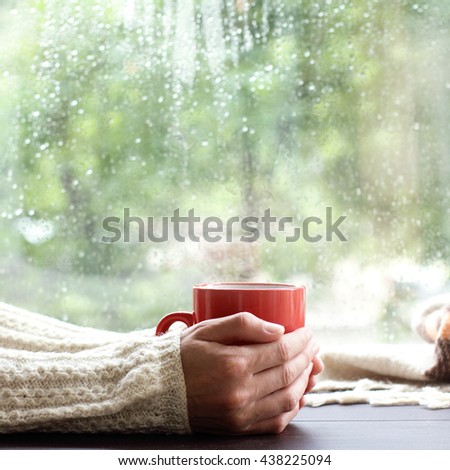 red mug with a hot drink in the hands of a person dressed in a warm sweater near the window / time to drink a warm drink