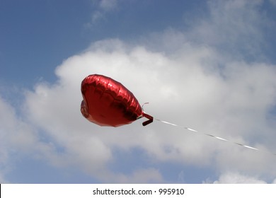 Red Mylar Heart Helium Balloon Floats Against The Blue Sky With White Fluffy Clouds