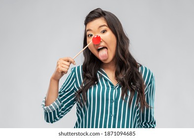 red nose day, party props and photo booth concept concept - happy asian young woman with clown nose sticking out tongue over grey background