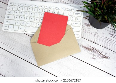 Red Note Inside Brown Envelope With Keyboard And Potted Plant Background On Wooden Desk. Copy Space. Flat Lay.