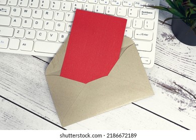 Red Note Inside Brown Envelope With Keyboard And Potted Plant Background On Wooden Desk. Copy Space. Flat Lay.