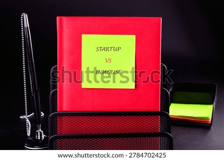 A red notebook with a yellow sticky note stating STARTUP VS INHOUSE is displayed in a black wire mesh organizer.