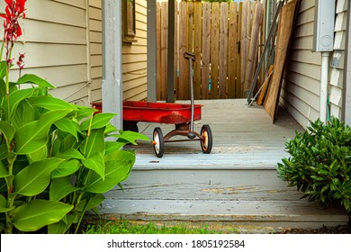 A red old rusty toy wagon was left on the doorway of an old house. The wooden porch the toy was left has grey paint which shows signs of aging. There are wooden fences on the other side of entrance.