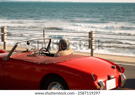 Red old timer car, the back of an elderly Japanese man drives a vintage red car along the scenic coastal roads of Kamakura in Japan with the blue ocean stretching out in the background with copy space