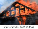 the red and orange flames hungrily lapping at the trusses of a house. the blackened timber of a building stands strongly against the angry flames surrounding it. Charred wood, flames and smoke against