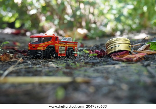 Red and orange toy truck in leaves next to a\
metal yellow lid on the ground. Photo on the background of green\
leaves and branches