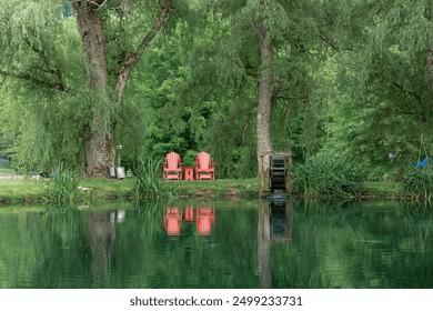 Red outdoor chairs and table in between two weeping willow trees in front of a pond. Reflection in water and a water mill to the side. Green trees, bushes and grasses everywhere.  - Powered by Shutterstock