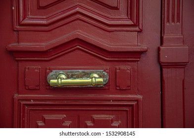 Red Painted Triangle Framed Wooden Door With Vintage Shiny Brass Handle On Marble Plate. Textured Surface Of Antique Door. Architectural Details Of Paris Door Of Old Classical Building In France.