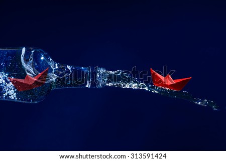 Red paper boat rides on a water splash out of the bottle, another waits for freedom inside at the bottleneck, dark blue background, concept for get away, escape or liberation