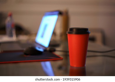 Red Paper Cup In The Office On The Table In The Evening With Lamp Light On The Background Of A Glowing Laptop. Work Space And Overtime.