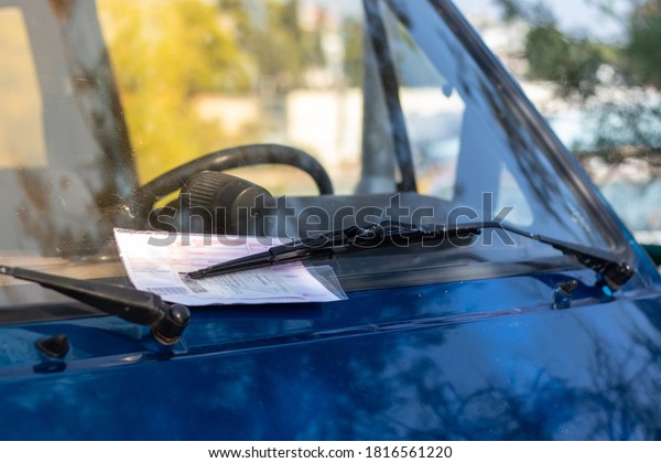 Red parking ticket stuck under the\
windshielf wiper of a blue car, person fined for illegal parking in\
public. Contents of the ticket blurred for\
privacy