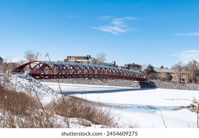 A red pedestrian bridge spans over a frozen river with a clear blue sky above. The banks are covered in snow, and bare trees can be seen in the distance, suggesting a cold winter day. - Powered by Shutterstock