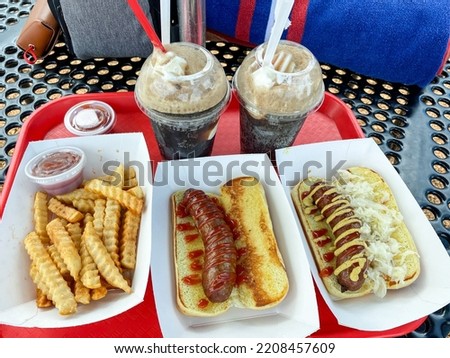 A red plactic tray has two root beer floats in plastic cups, with three white trays:containing french fries, one brat with ketchup and one brat with ketchup, mustard, and sauerkraut on toasted buns.
