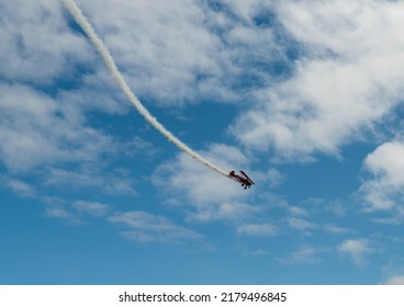 A Red Plane Is Flying Down Leaving A White Smoke Trail Behind It And Showing Aerobatics In A Blue Sky With Clouds