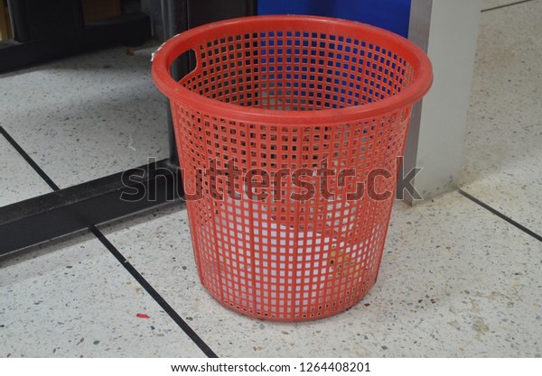 Red plastic basket adapted to be used as garbage\
bin containing sheet of paper for reuse, put on a marble tiled\
floor in a corner of an\
office