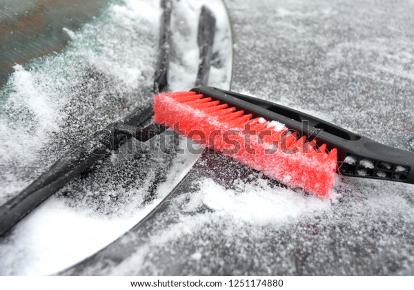 Red plastic\
brush cleaning snow. Shoveling snow from automobile. Grey car\
outside covered with snow and ice. Frozen window glass with\
selective focus at the winter blizzard\
day.